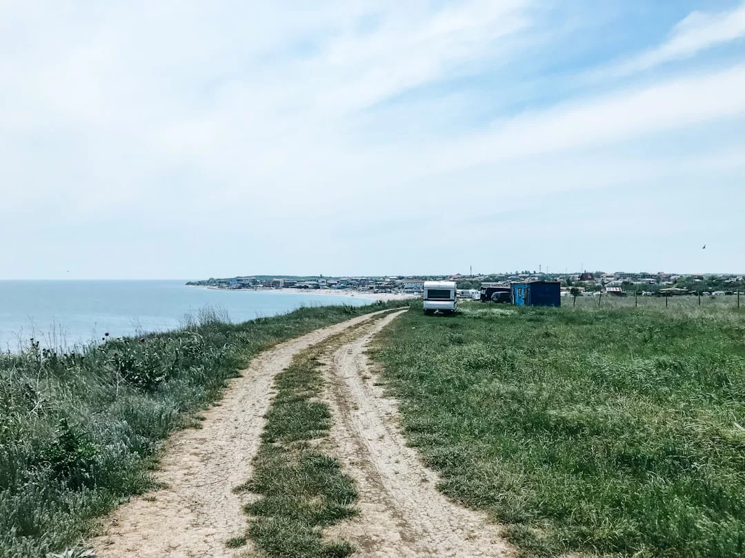 A dirt road curves along a grassy field beside the sea, leading to a parked van and a blue structure—an inviting scene that hints at unique real estate opportunities, with a distant town visible under a partly cloudy sky.