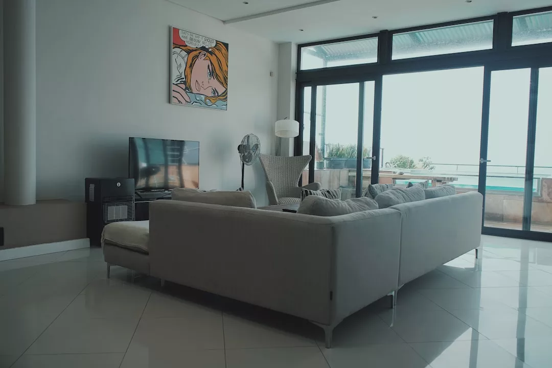 Modern living room with a gray sectional sofa, TV, fan, and large windows facing a balcony; contemporary artwork hangs on the wall above the TV—perfect for showcasing stylish real estate.