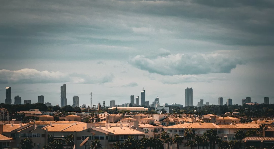 A city skyline with tall buildings in the distance under a cloudy sky, and tan-roofed residential houses in the foreground—perfect for real estate seekers exploring vibrant urban living options.