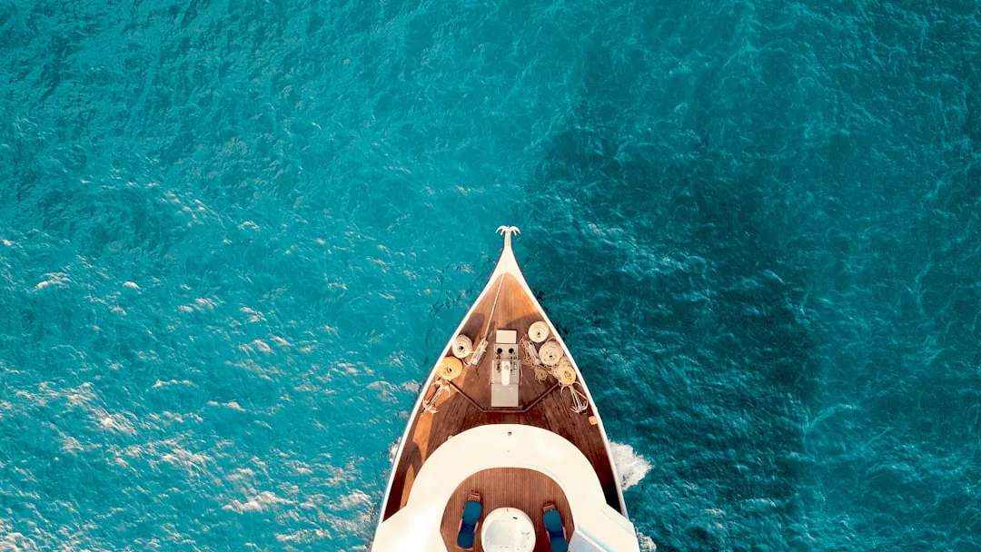 Aerial view of a white yacht sailing on clear blue water, highlighting the boat’s bow, deck, and surrounding sea—a scene of luxury perfect for exclusive real estate marketing.