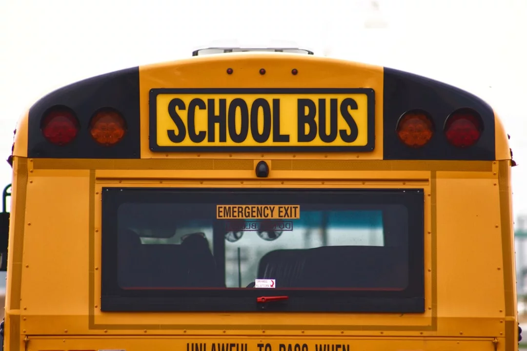 The image shows the rear view of a yellow school bus with "SCHOOL BUS" and "EMERGENCY EXIT" signs visible above and below the rear window, parked near a residential area—highlighting neighborhood amenities for real estate listings.