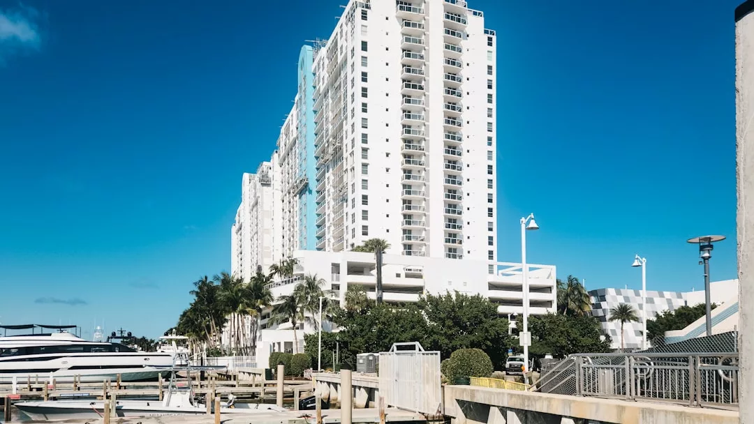 A tall white apartment building with balconies stands near the marina, offering prime real estate surrounded by boats and palm trees under a clear blue sky.