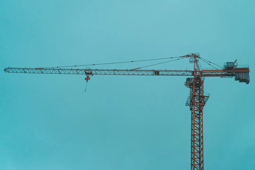 A tall, orange construction crane stands against a clear, blue sky, its long horizontal arm extending to the left—signaling new Real Estate development on the rise.