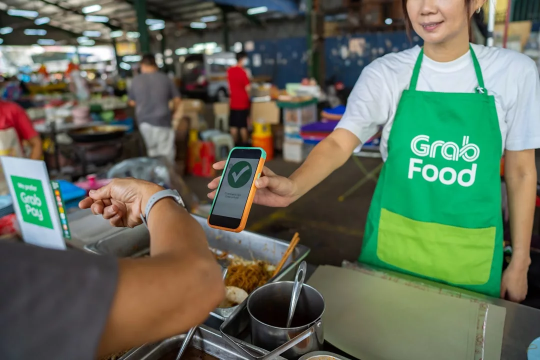 A vendor in a GrabFood apron shows a confirmation screen on a smartphone to a customer at a food market stall, indicating successful mobile payment—demonstrating how digital transactions extend beyond Real Estate into everyday experiences.
