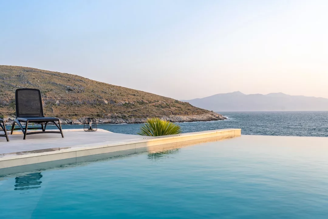 A modern infinity pool overlooks the ocean, enhancing this stunning real estate with a lounge chair on the deck and hills in the background under a clear sky.