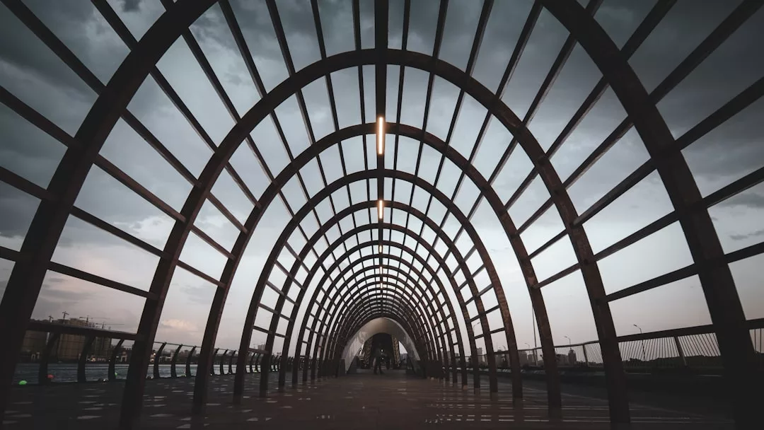 Arched metal pedestrian bridge with geometric grid pattern, photographed from underneath at dusk with a cloudy sky in the background—an architectural feature that adds modern appeal to surrounding real estate.