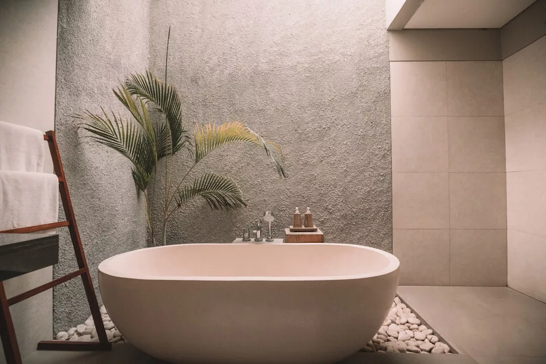 Modern bathroom with a freestanding bathtub on smooth pebbles, a textured wall, potted plant, and towels on a wooden rack—perfect for adding spa-like luxury to your real estate listing.
