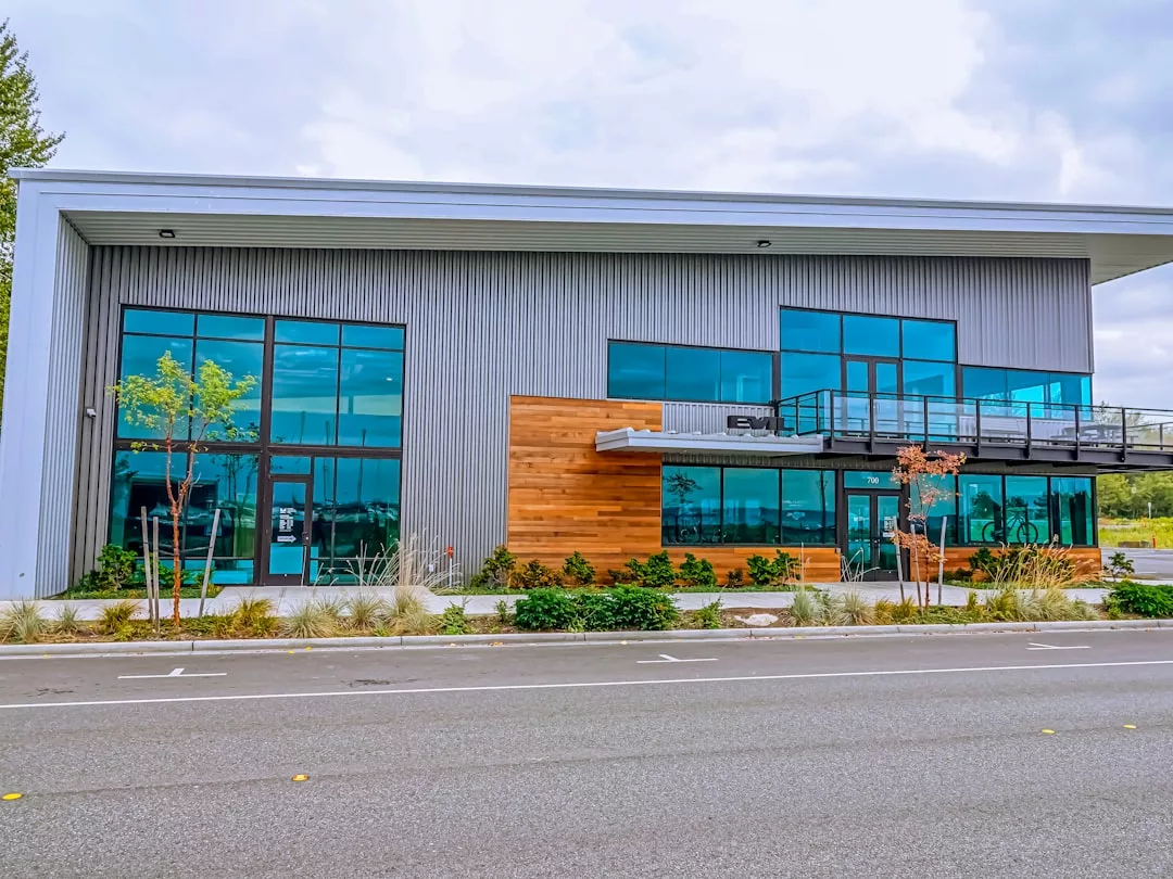 Modern two-story commercial real estate building with large blue-tinted windows, metal siding, wood accents, and a balcony, viewed from across an empty street.