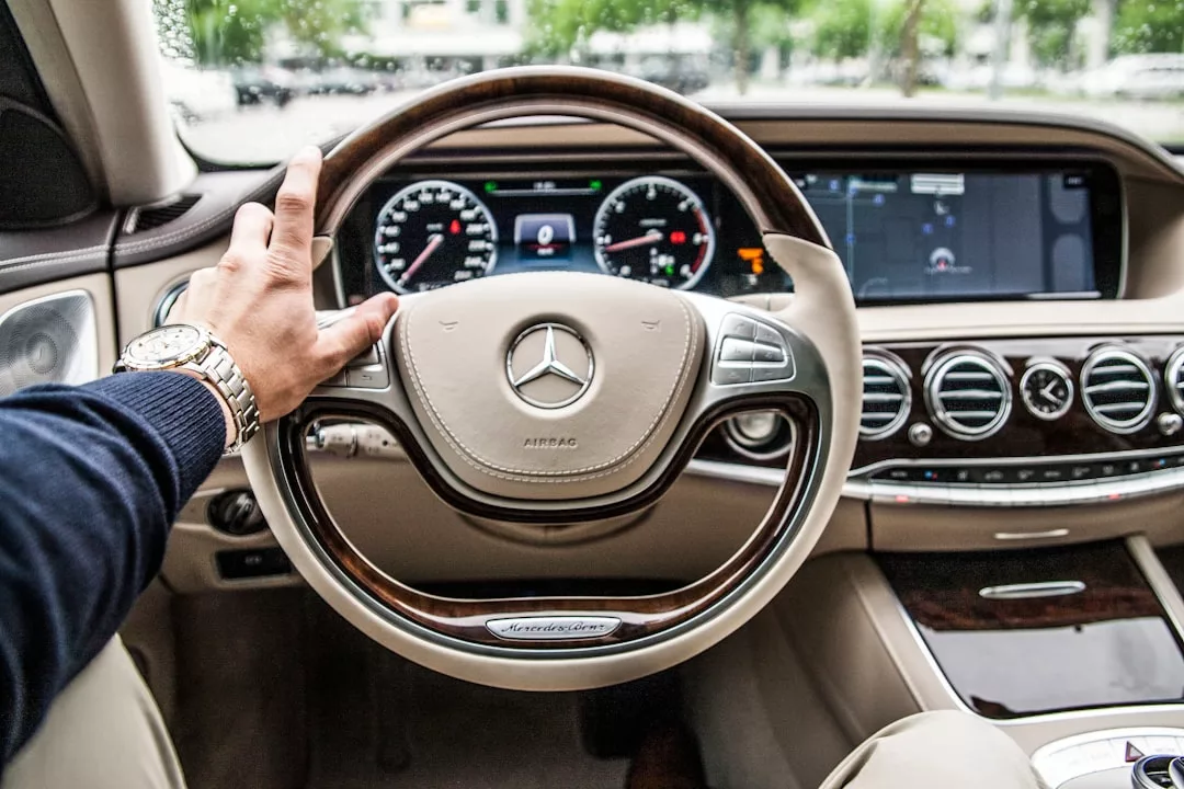 A person’s left hand on the steering wheel of a Mercedes-Benz car, showing the dashboard, speedometer, and infotainment screens—capturing a moment where luxury vehicles meet real estate success.