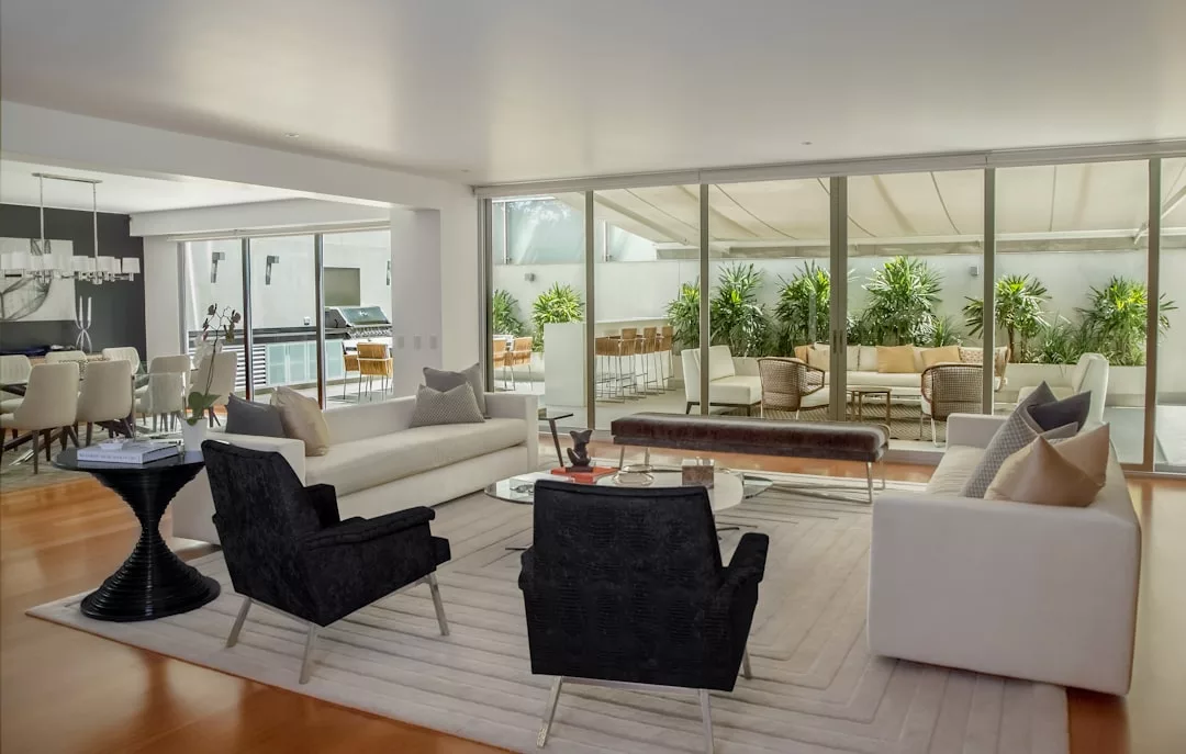 Modern living room with white sofas, black armchairs, and a glass coffee table, featuring large sliding glass doors opening to a patio with greenery—an inviting space perfect for real estate showings. Dining area visible in the background.