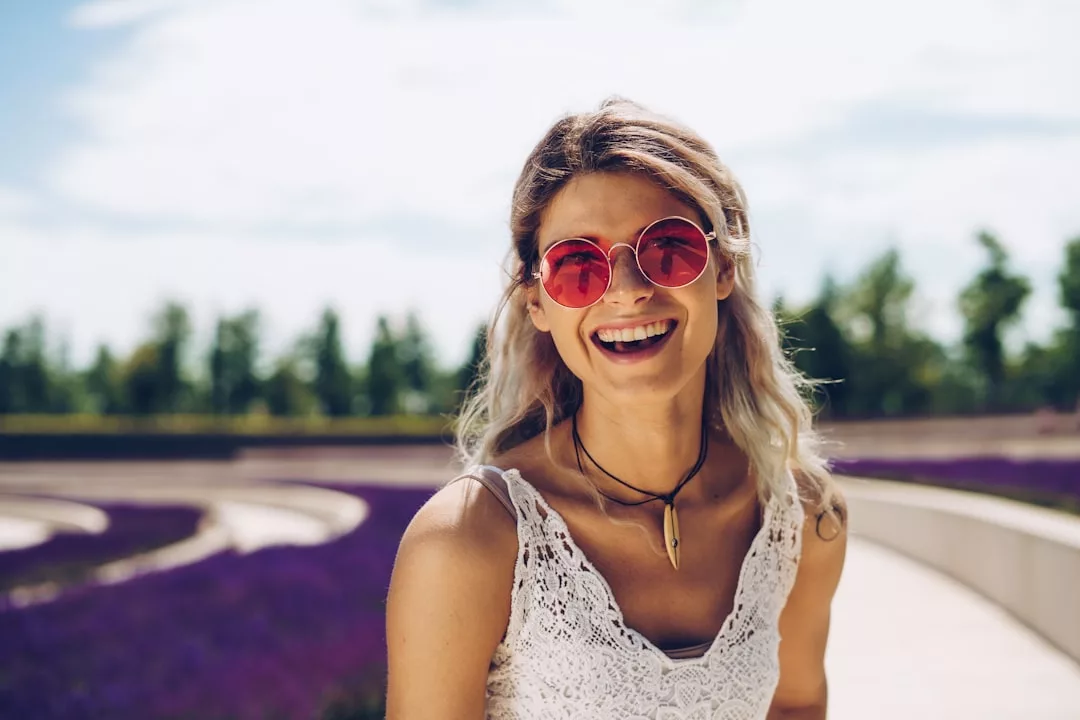 Smiling woman wearing pink sunglasses and a white lace top stands outdoors near curved pathways and a field of purple flowers, showcasing the serene landscape perfect for real estate opportunities, with trees and sky in the background.