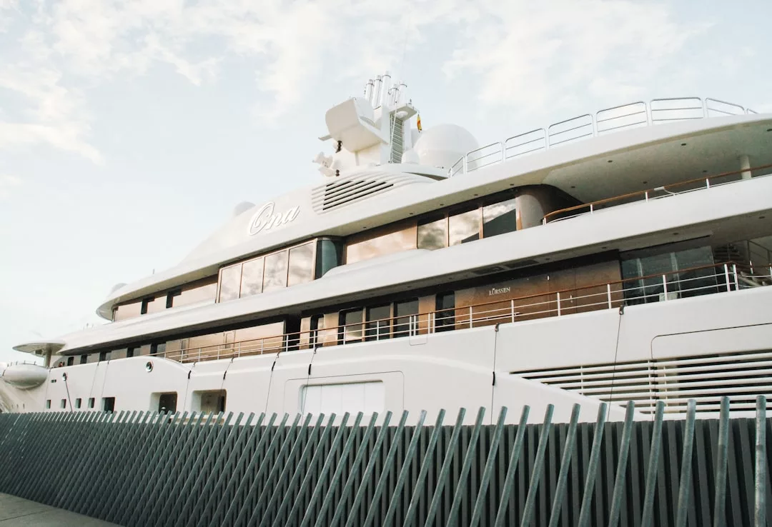 A large white luxury yacht, reminiscent of real estate grandeur, docked at a marina and viewed from the side behind a metal fence under a partly cloudy sky.
