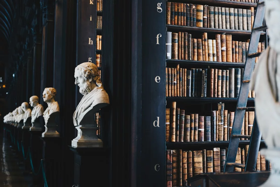 Marble busts line a hallway beside tall bookshelves filled with old books and a wooden ladder in this historic library—a timeless gem for real estate enthusiasts seeking classic elegance.