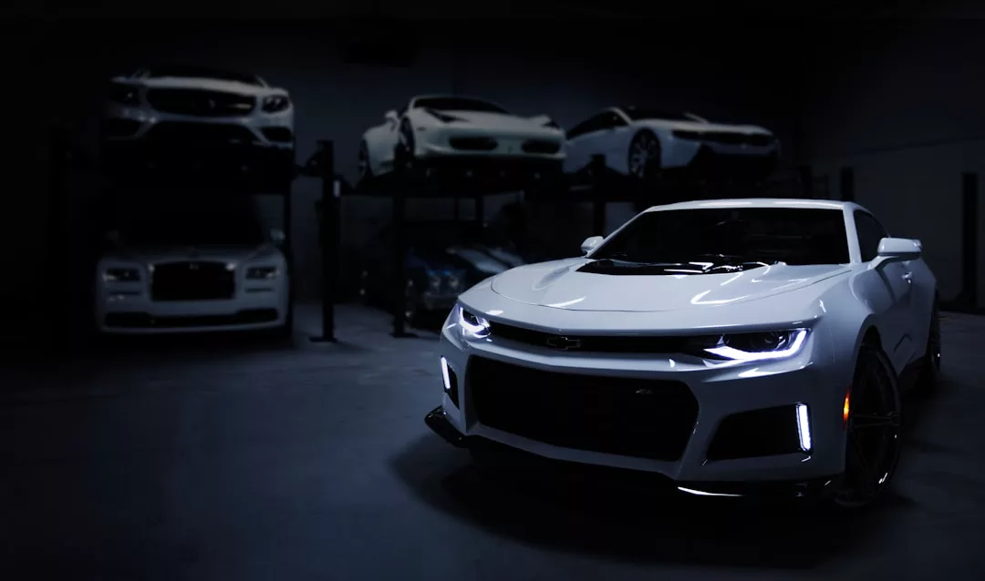 A white Chevrolet Camaro is parked in a dimly lit garage, with several luxury cars visible on lifts in the background—a scene reflecting the ultimate dream for real estate enthusiasts who appreciate high-end living and premium vehicles.