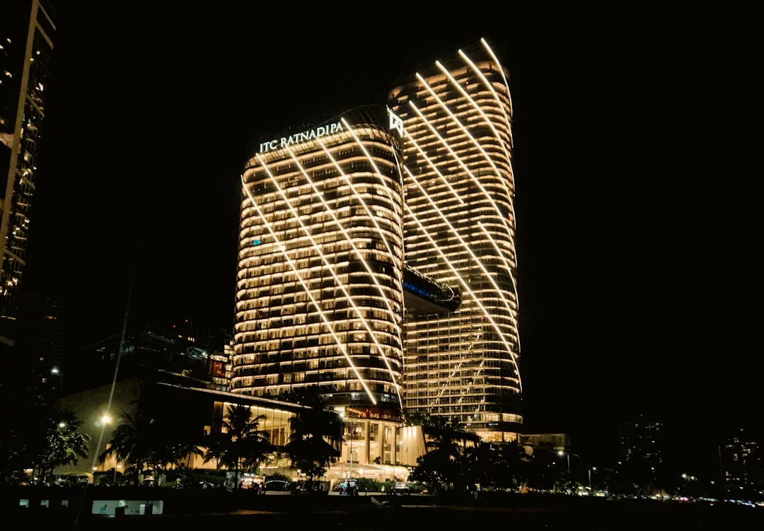 Two modern, high-rise Real Estate buildings with "ITC Ratnadipa" signage are illuminated by diagonal lights at night, with palm trees and a few cars visible at the base.