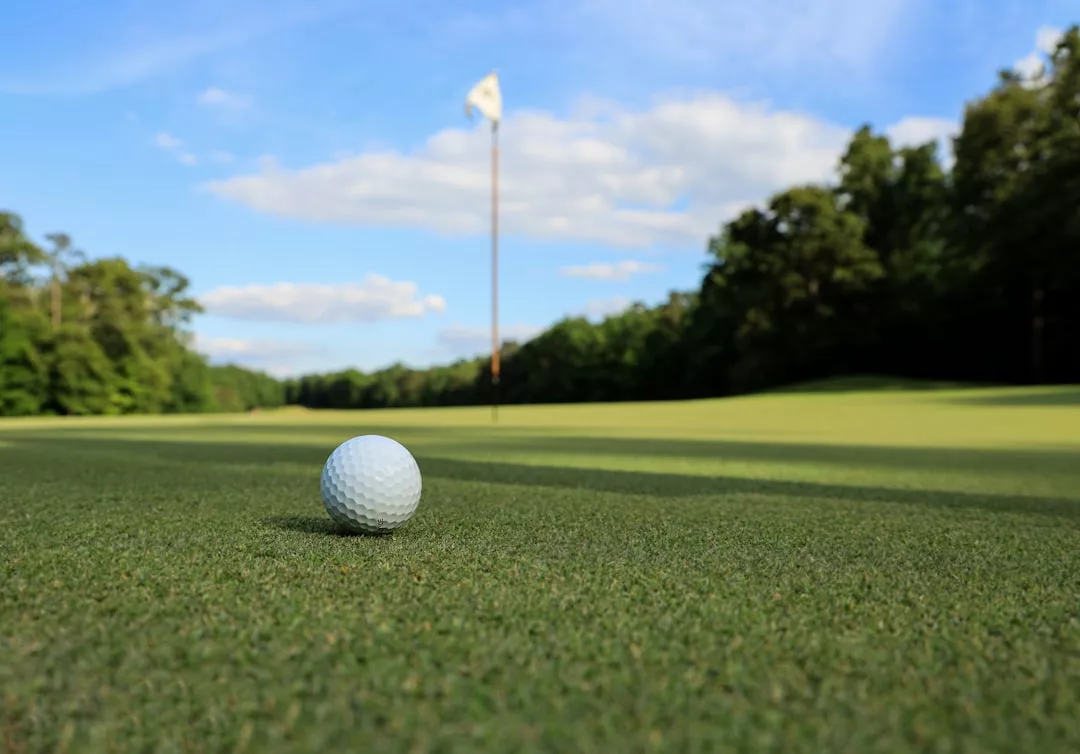 A golf ball rests on the green with a flagstick and hole visible in the distance under a partly cloudy sky, evoking the serenity often sought in prime real estate locations.