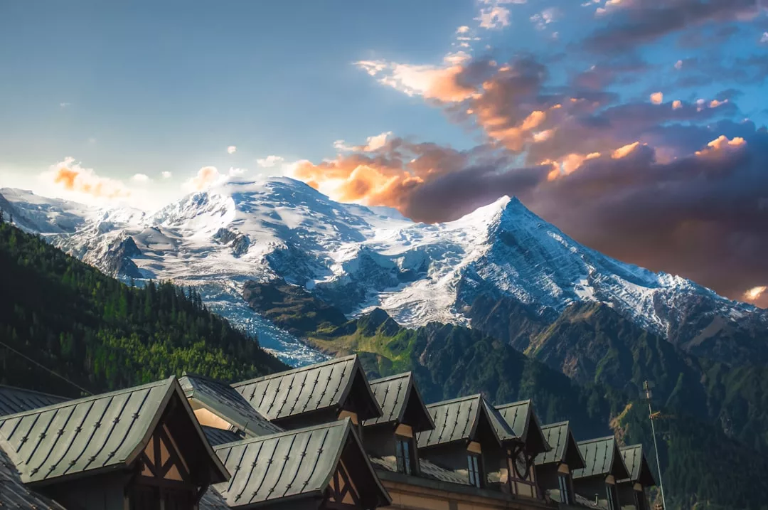 Snow-capped mountains under a dramatic, colorful sky overlook rows of rooftops in the foreground, offering a unique real estate setting surrounded by green forested slopes below the peaks.