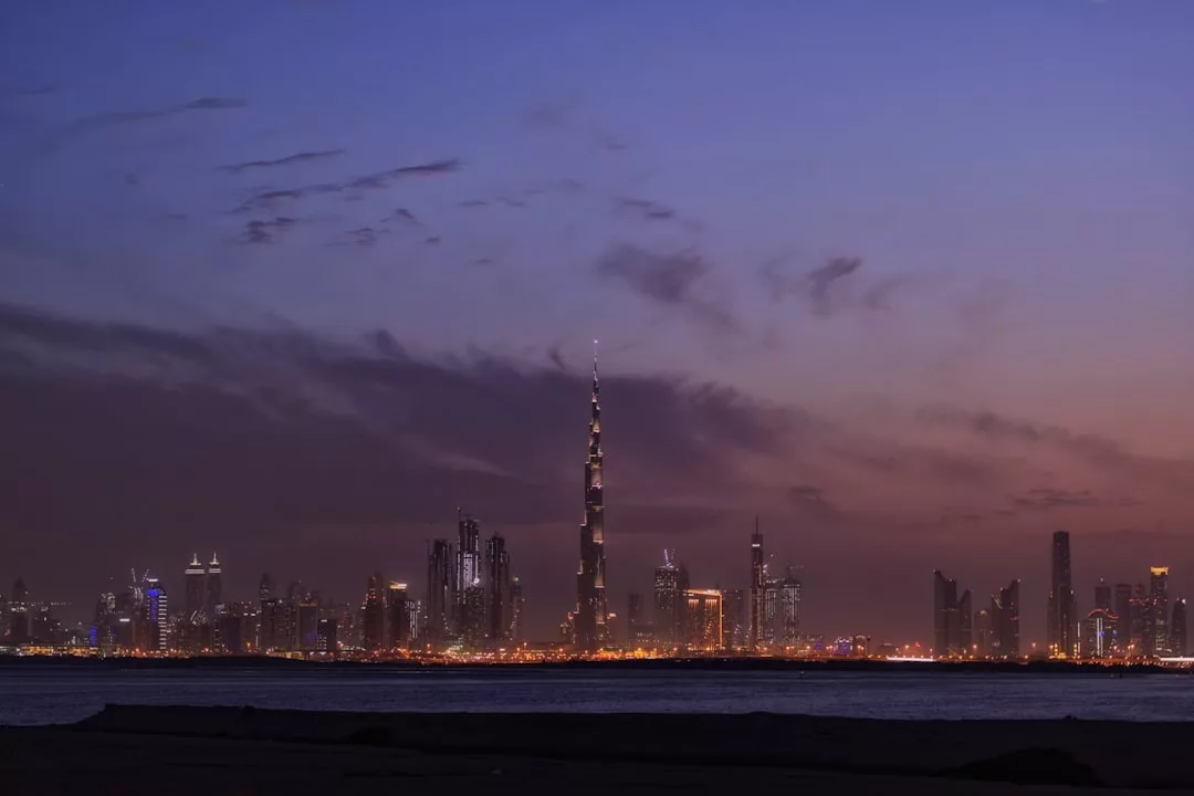 City skyline at dusk with illuminated buildings and a prominent tall tower in the center, showcasing prime real estate, viewed from across a body of water under a partly cloudy sky.