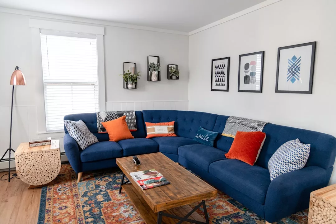A stylish living room perfect for real estate showcases, featuring a blue sectional sofa, colorful pillows, a wooden coffee table, patterned rug, wall art, and plants on bright white walls.