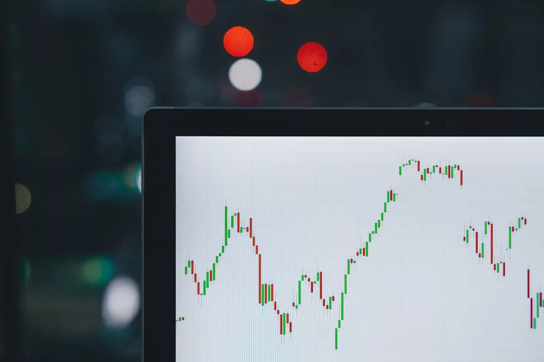 A computer screen displays a fluctuating financial chart with red and green candlesticks, reflecting Copenhagen property prices; blurred lights appear in the background.