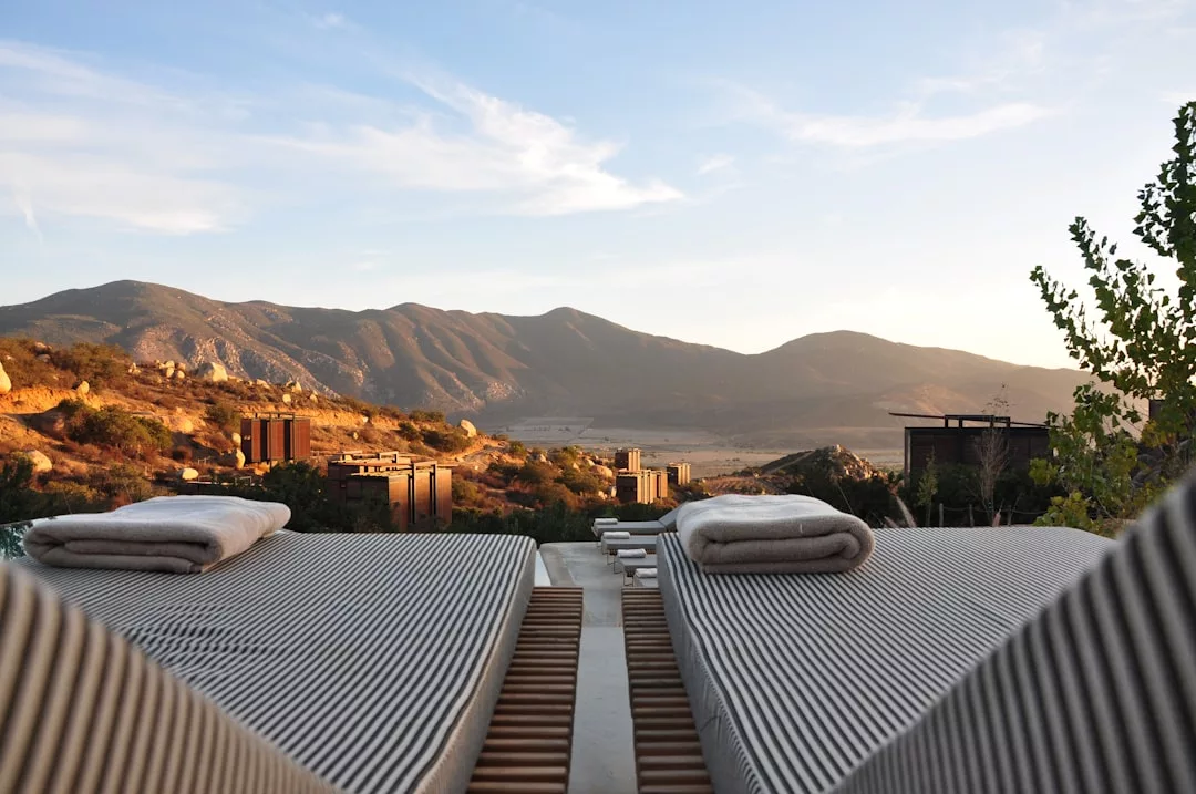 Two lounge chairs with towels face a scenic mountain landscape at sunset, overlooking several modern real estate buildings scattered in the valley below.
