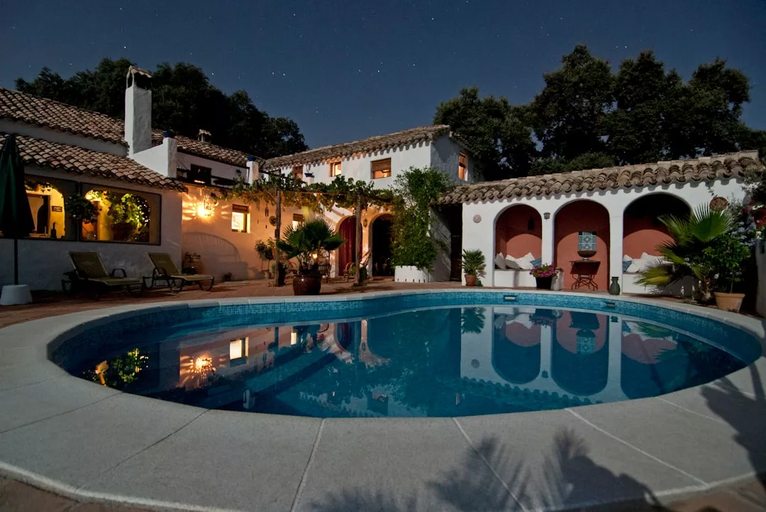A Mediterranean-style real estate villa with a tiled roof and arched patio surrounds a lit outdoor swimming pool at night.