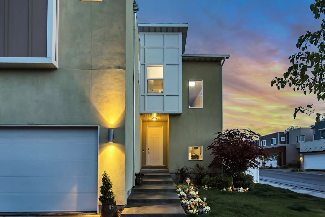 Modern two-story house perfect for real estate buyers, with lights on, a white garage door, small front garden, and a walkway leading to the main entrance at sunset.