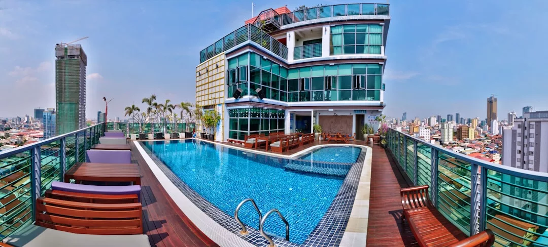Rooftop pool with wooden deck, lounge chairs, and glass-railed fencing on a modern building—an exceptional real estate gem overlooking the city skyline on a clear day.