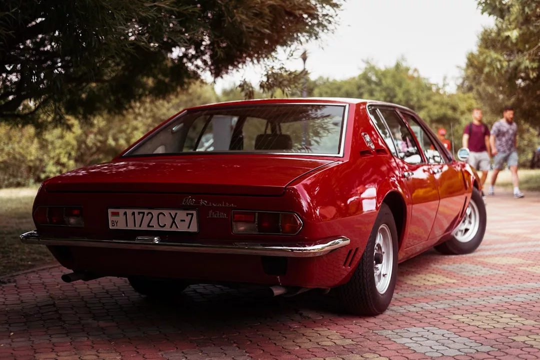 A red classic Alfa Romeo car is parked on a paved pathway, adding vintage charm to this real estate setting, while two people stroll in the background among the trees.
