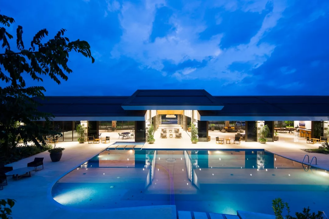 Modern real estate building with large open spaces and glass walls, illuminated at dusk, features a central swimming pool reflecting the lights and blue sky.