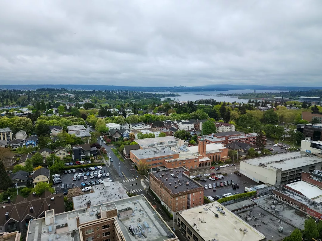 Aerial view of a suburban Real Estate area with residential buildings, parking lots, and trees under a cloudy sky, with a body of water visible in the distance.