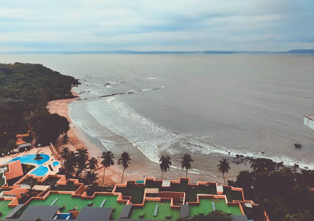 A coastal real estate resort with a pool and palm trees sits beside a sandy beach, where waves approach the shore under a cloudy sky.
