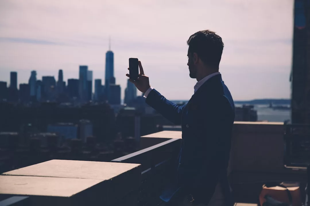 A man in a suit stands on a rooftop terrace, taking a selfie with his smartphone against the city skyline, showcasing the allure of urban real estate.