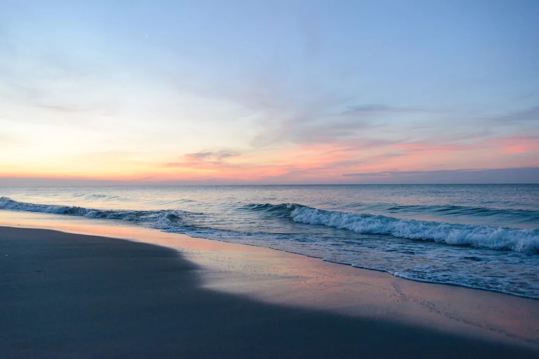 Calm ocean waves wash onto a sandy beach at sunset, pastel colors reflecting on the water—an idyllic setting perfect for luxurious real estate and coastal living.