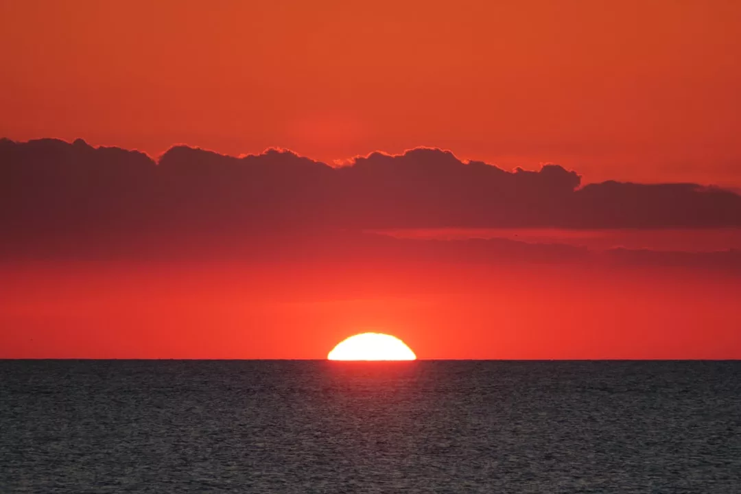 The sun is setting over the ocean, partially hidden by the horizon, with dark clouds and an orange-red sky above—an inspiring view for prime real estate along the coast.