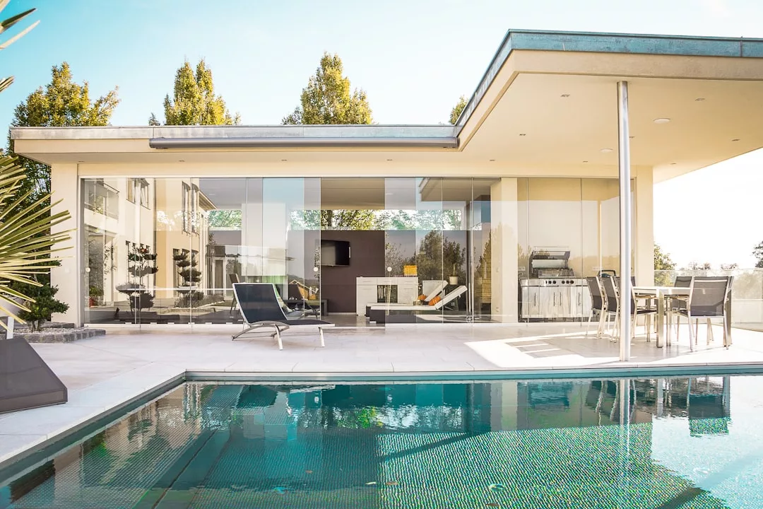 Modern real estate gem with large glass windows, poolside patio, lounge chairs, outdoor dining area, and barbecue grill, surrounded by trees on a sunny day.