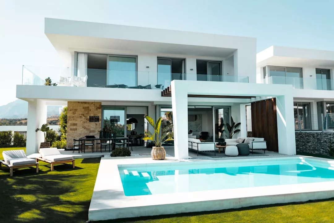 Modern two-story white house with large windows, a balcony, outdoor seating area, swimming pool, and landscaped lawn under clear skies—a stunning real estate opportunity.
