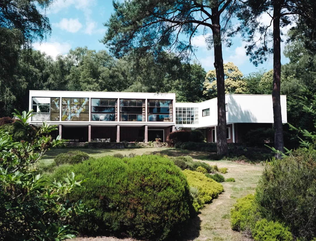 A modernist two-story house with large glass windows sits among trees and landscaped greenery under a partly cloudy sky, offering an impressive real estate opportunity.